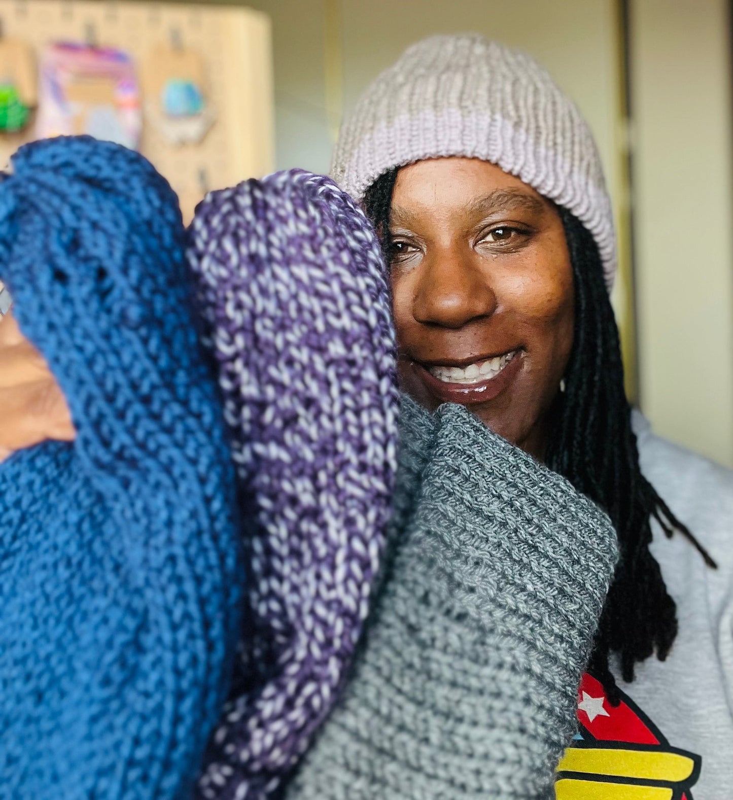 Tara is smiling while wearing a beige knit beanie and holding a stack of 3 beanies.